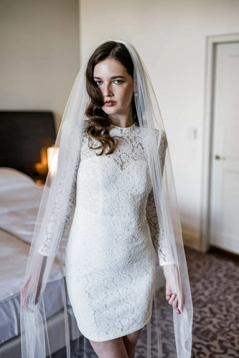 Braut steht mit Schleier und Spitzenkleid im Hotelzimmer