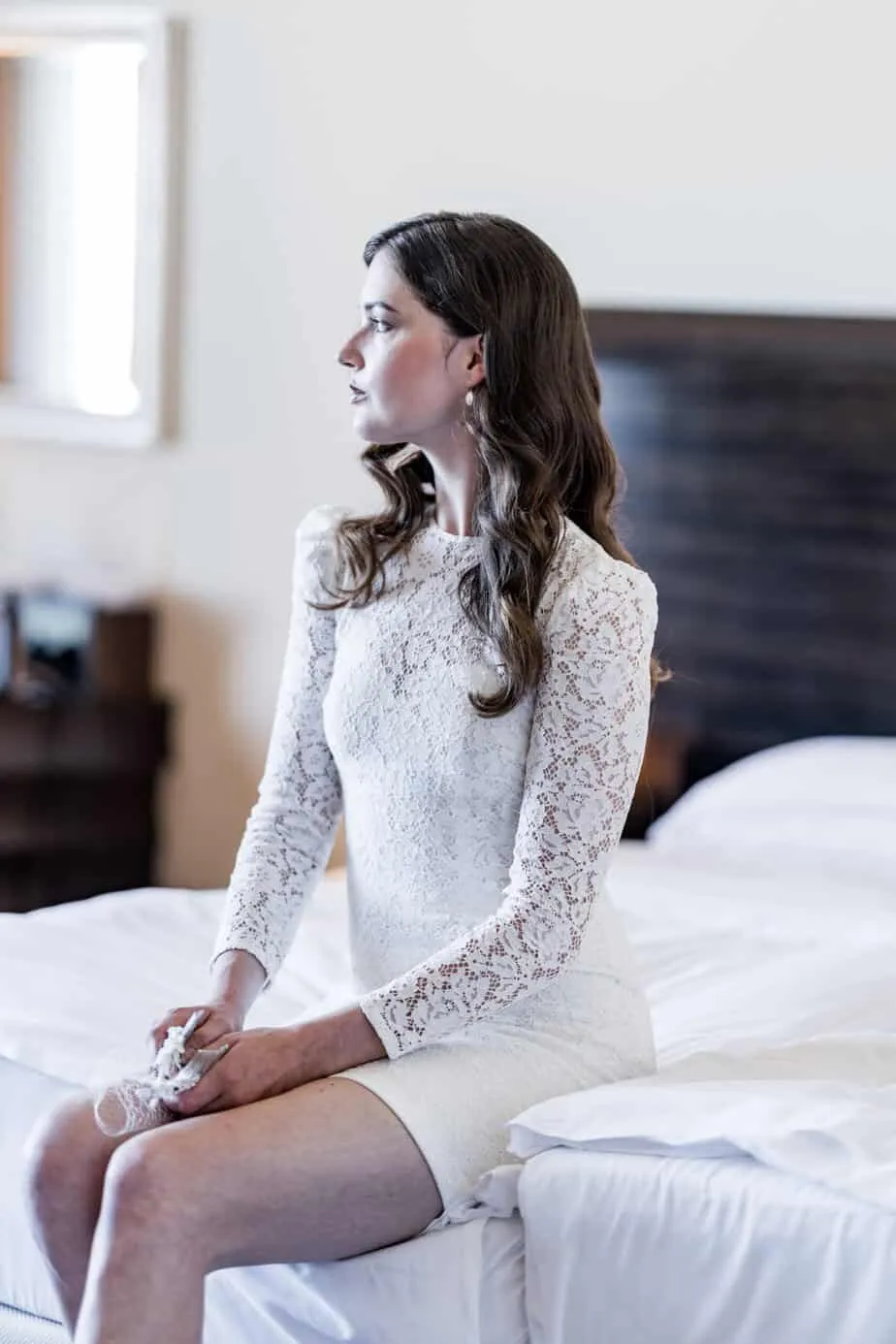 Braut sitzt im Spitzenkleid auf dem Bett ihrer Hotelsuite