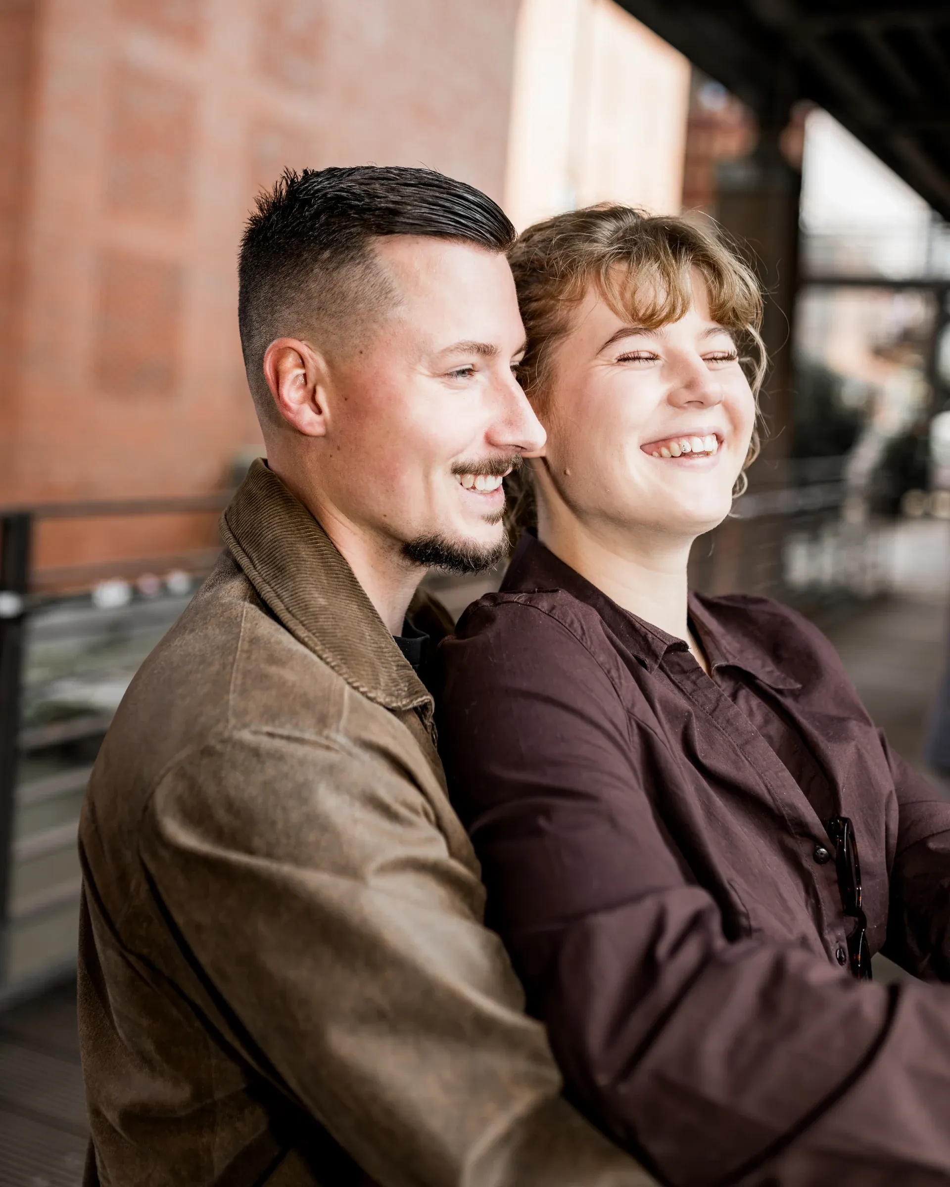 Liza und Domenic sitzen lachend vor einer Backsteinfassade