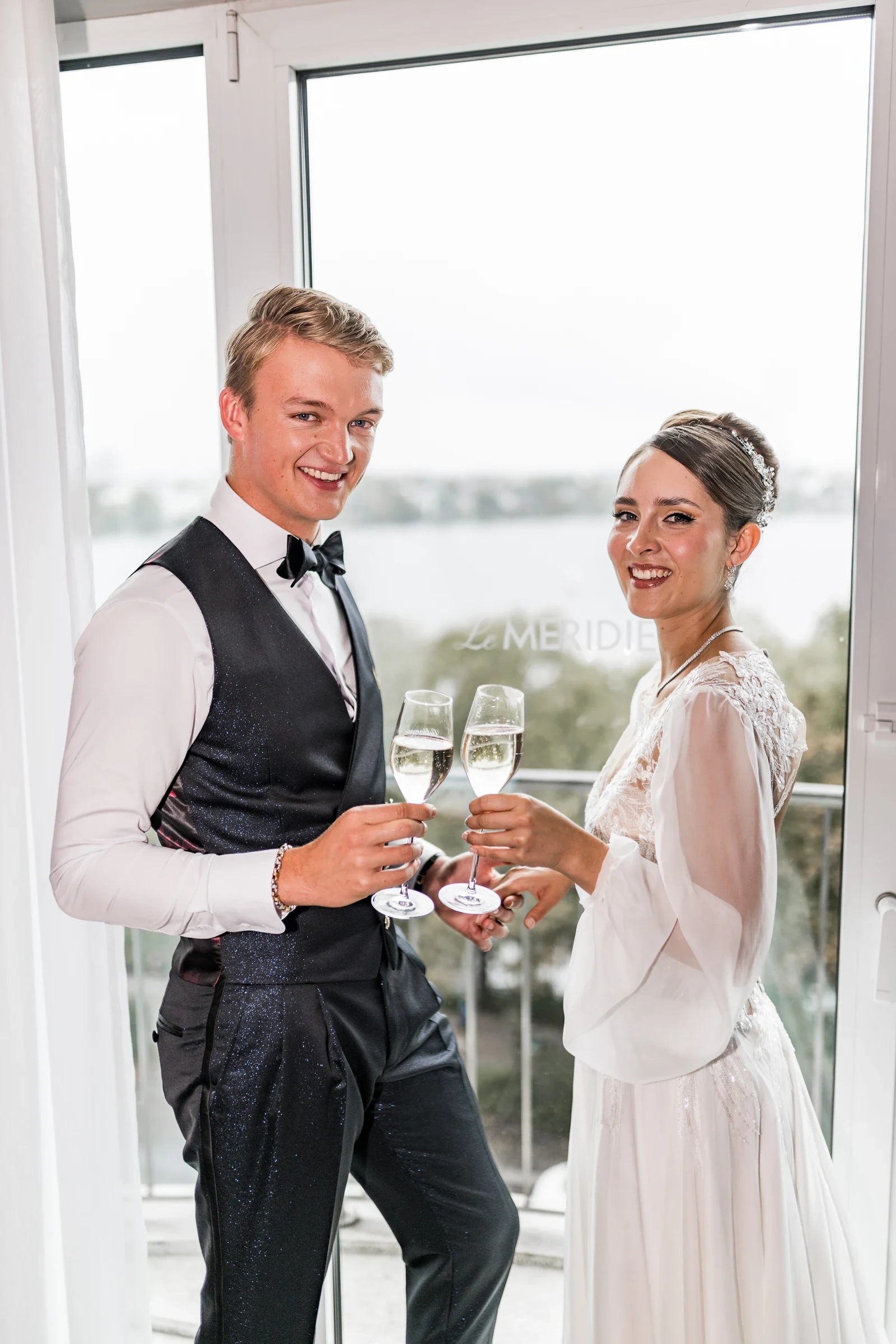 Junges Brautpaar in festlicher Hochzeitstracht stößt mit Sektgläsern vor einem großen Fenster mit Seeblick an.