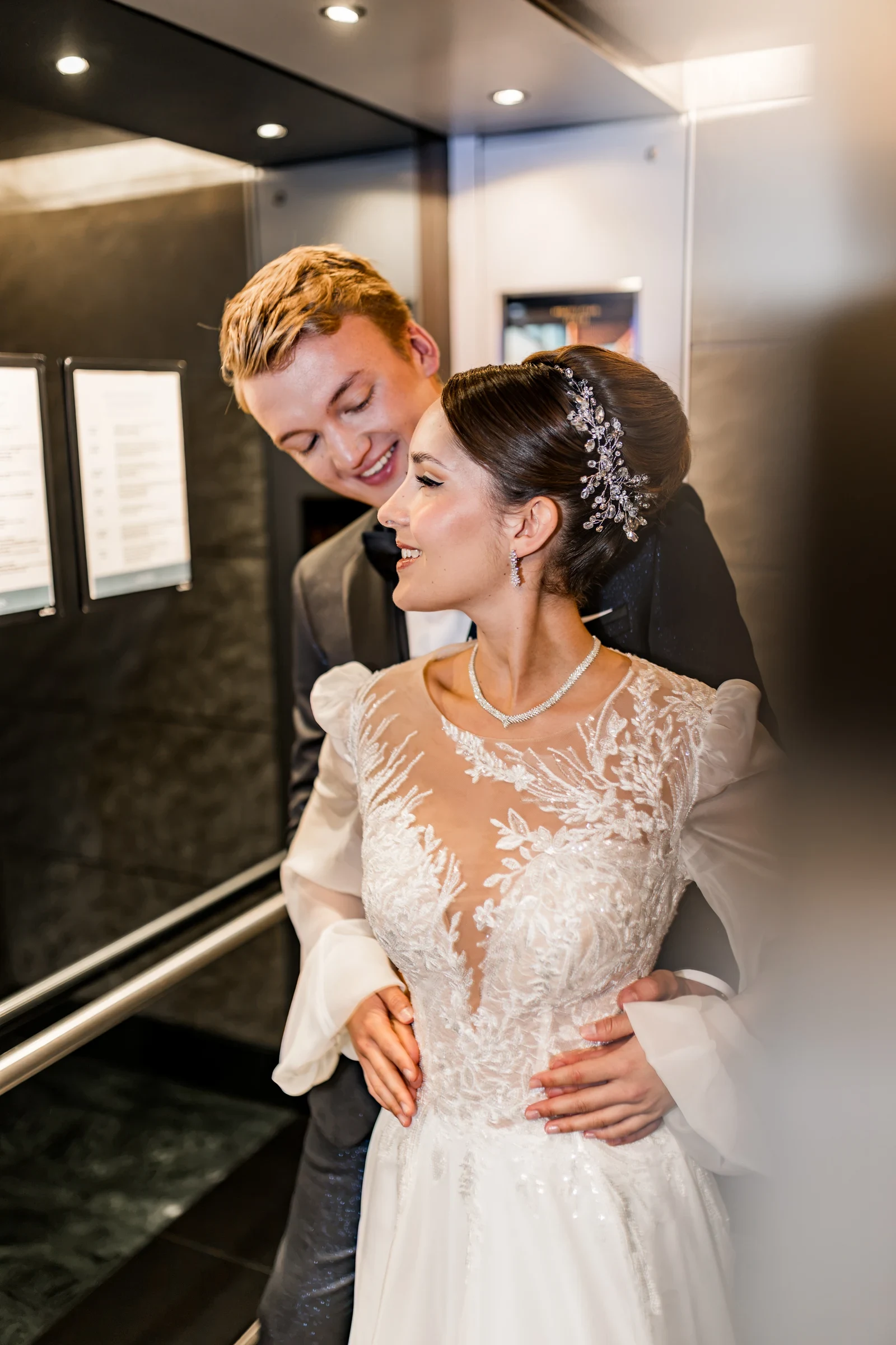 Braut und Bräutigam umarmen sich innig in einem Aufzug, die Braut trägt ein detailliertes Spitzen-Brautkleid mit transparentem Oberteil und Schmuck im Haar.