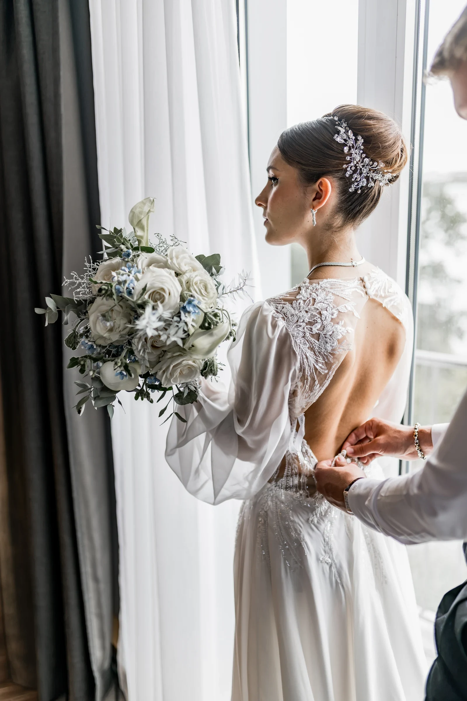 Braut im weißen Spitzenbrautkleid mit Hochsteckfrisur hält einen großen Brautstrauß und wird von einer Person am Rücken des Kleides zugeknöpft.