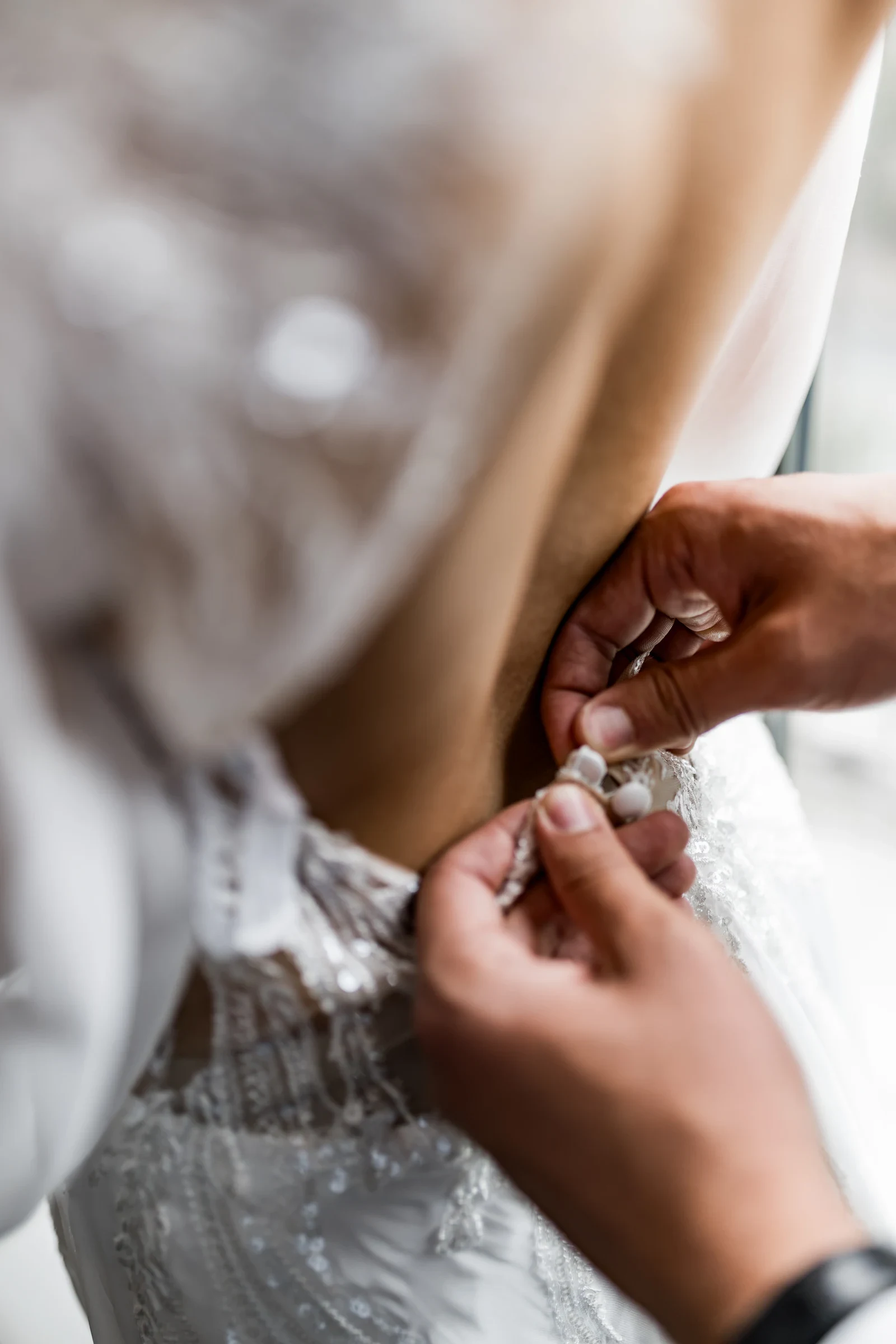 Hände schließen feine Knöpfe am Rückenteil eines bestickten Brautkleids während einer Hochzeitsvorbereitung