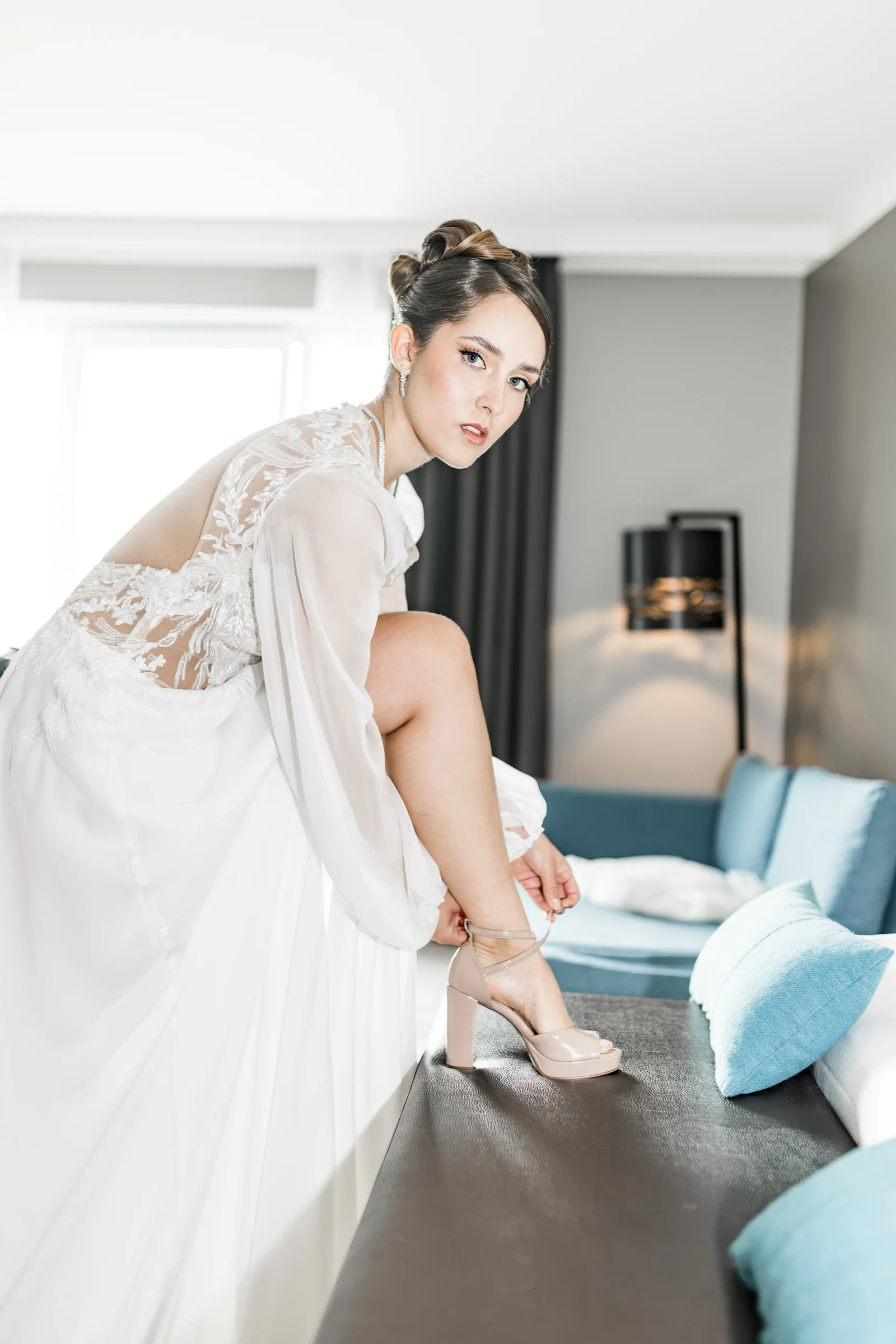 Braut in einem eleganten, weißen Brautkleid mit transparenten Spitzenärmeln sitzt auf einer Bank und zieht sich beige High-Heel-Sandalen an, im Hintergrund ein modernes Wohnzimmer mit hellen Kissen und Lampe.