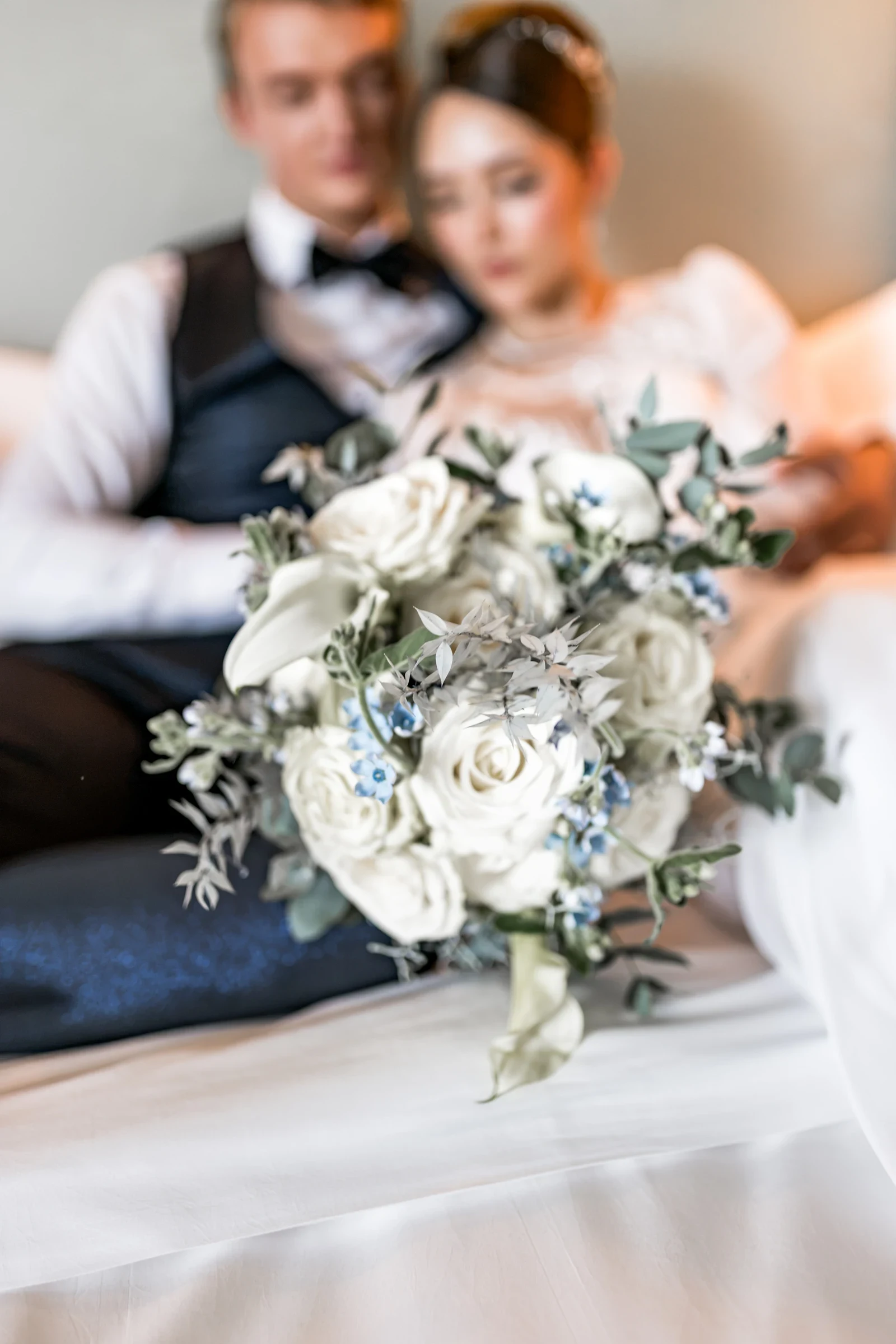 Brautpaar lässig auf einem Bett sitzend, im Vordergrund ein strahlender Brautstrauß mit weißen Rosen und blauen Akzenten