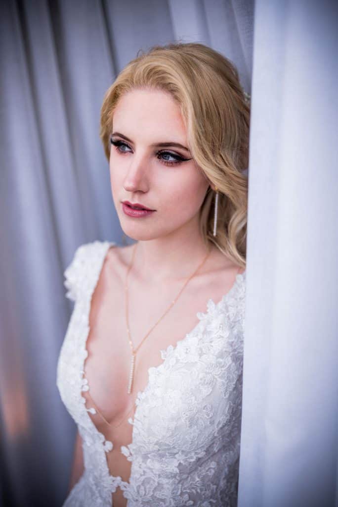 Blonde Braut in einem Spitzenhochzeitskleid posiert elegant vor einem Vorhang und präsentiert ihr Make-up und ihre Frisur.