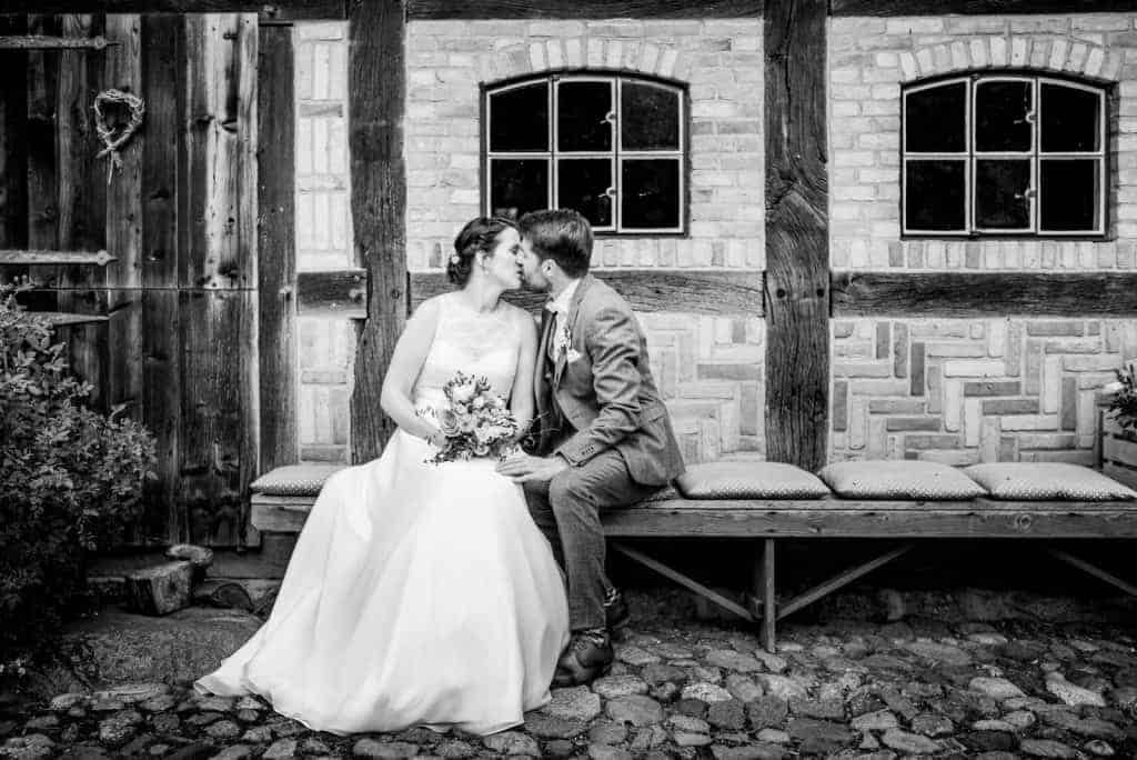 küssendes Brautpaar sitzt auf einer Bank vor einem Fachwerkhaus
