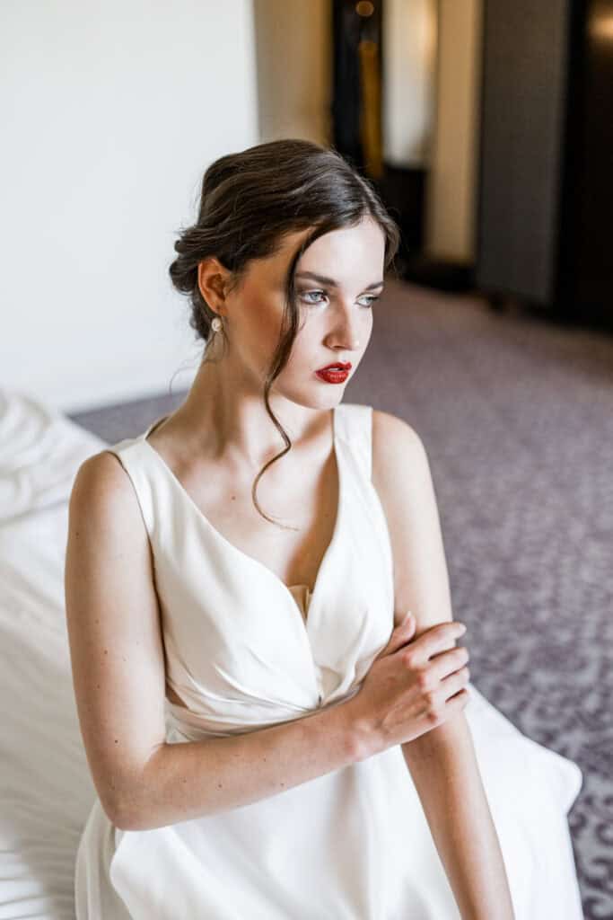 Elegante Braut in weißem Spitzenkleid mit roten Lippen und elegantem Haarstyling, sitzt nach der Hochzeit im Hochzeitszimmer, exklusive Hochzeitsfotografie.