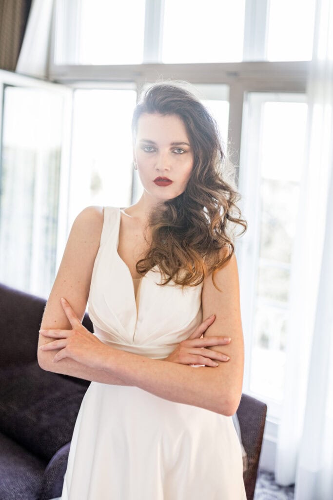 Elegante Frau in einem weißen Kleid, stehend in einem hellen Raum mit großen Fenstern, modernem Ambiente, professionelle Hochzeits- oder Modefotografie, natürlichen Licht-Effekt, Fokus auf Gesicht und Haar.