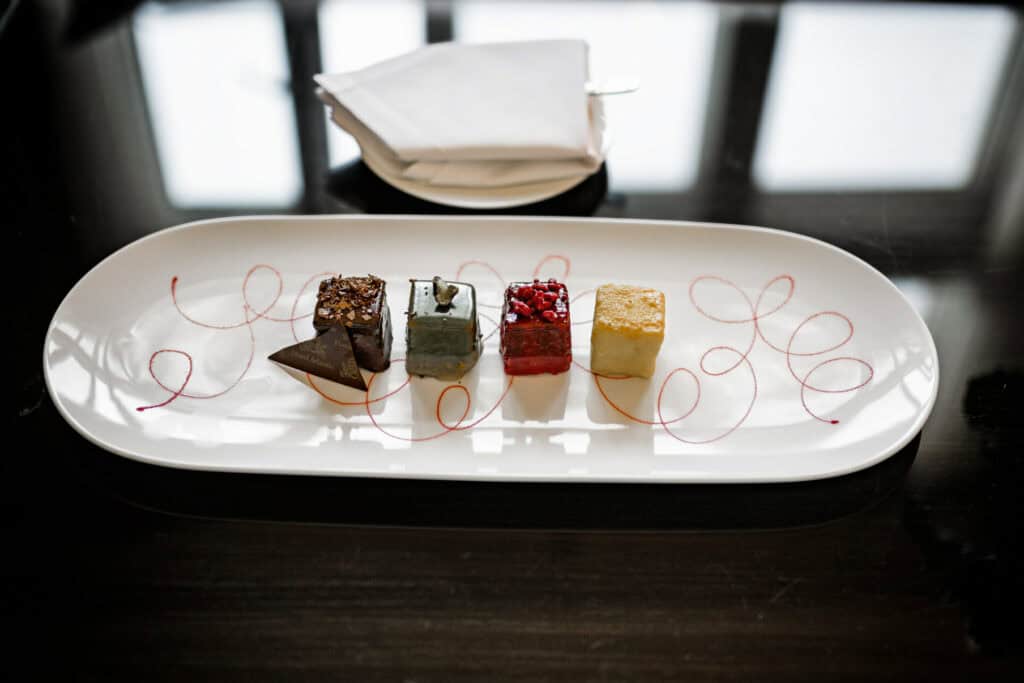 Verschiedene Gourmet-Desserts auf einem weißen Teller mit dekorativen Soßenwirbeln auf einem dunklen Tisch.