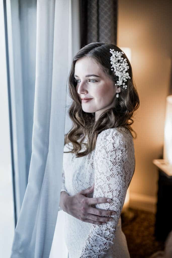 Elegante Braut in weißem Spitzenkleid mit Haaraccessoire, blickt verträumt aus dem Fenster, innerer Moment der Vorfreude vor der Hochzeit.