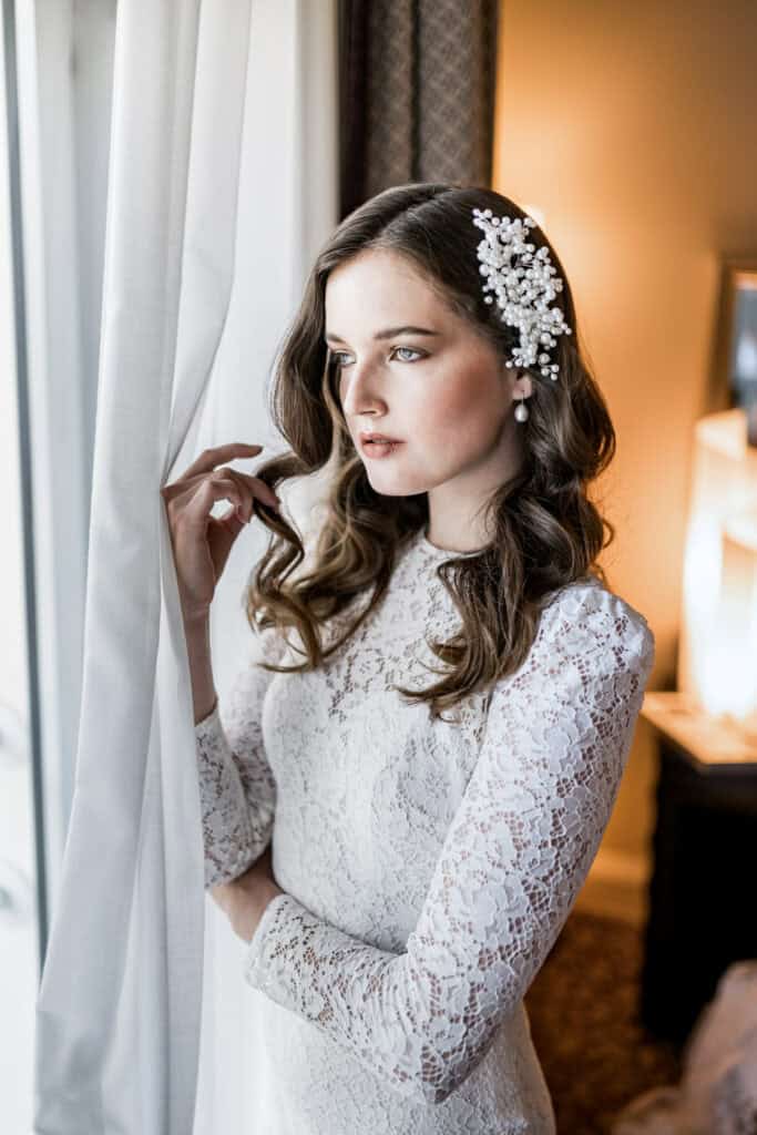 Feierliche Braut in eleganter Lace-Hochzeitskleidung mit Perlenhaarschmuck, blickt nachdenklich am Fenster.