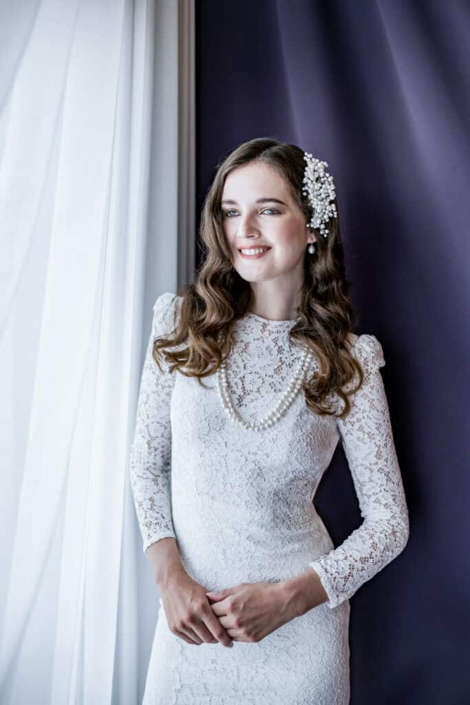 Elegantes Brautporträt mit weißem Spitzenkleid, Perlenkette und Blumenschmuck in den Haaren, natürlicher Lichtschatten am Fenster, stilvolle Hochzeitsfotografie, emotionale Atmosphäre, Hochzeitsfotografie, Porträtfotografie, Architekturlichteffekte, romantischer Moment.