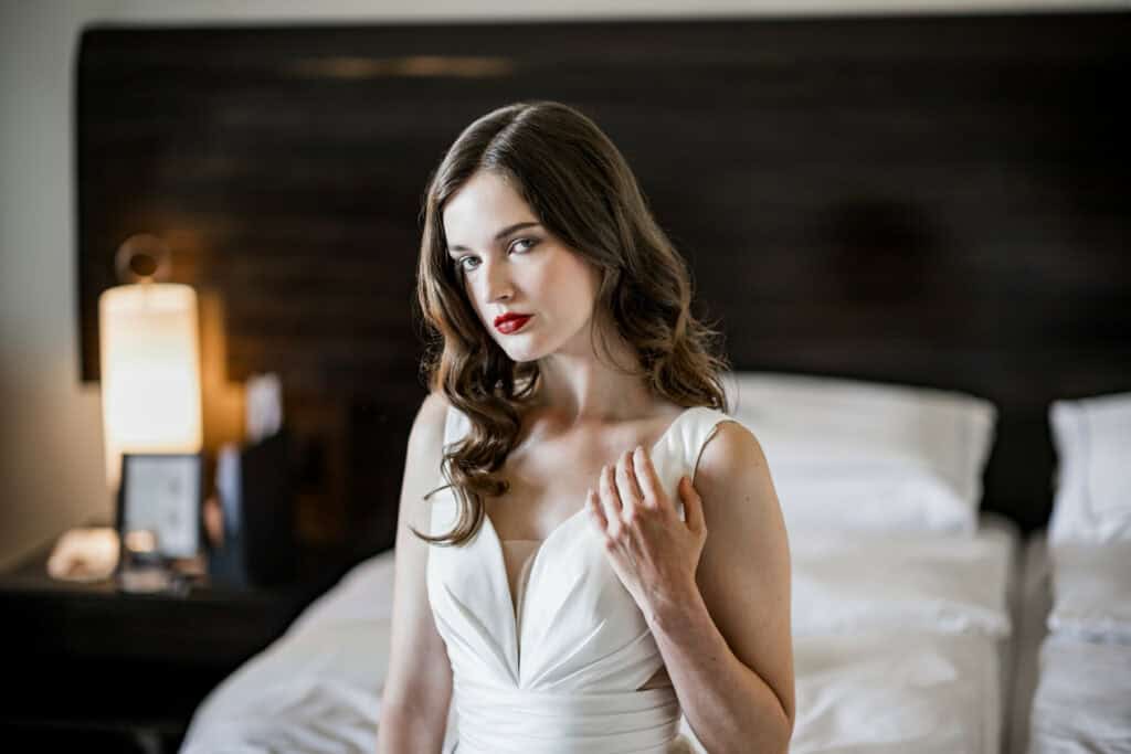 Elegante junge Frau im weißen Brautkleid in einem modernen Hotelzimmer, makelloser Look, schöne Locken, roter Lippenstift, stilvoller Hochzeitslook, Hochzeitsfotografie, Modefotografie, professionelle Portraitaufnahme.