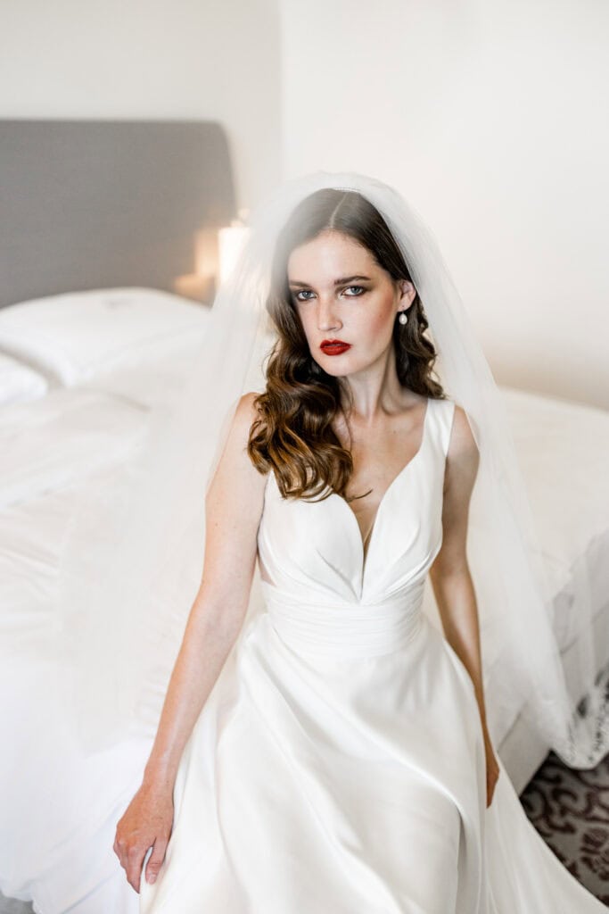 Elegante Braut in weißem Hochzeitskleid mit Schleier auf einer weißen Bett in moderner Hotelzimmer-Atmosphäre.