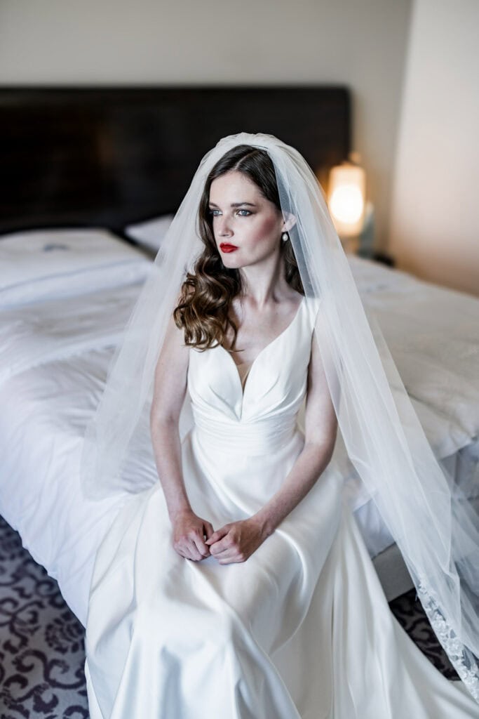 Elegante Braut im weißen Hochzeitskleid mit Schleier, sitzend auf dem Bett in einem modernen Hotelzimmer, bereit für den großen Tag.