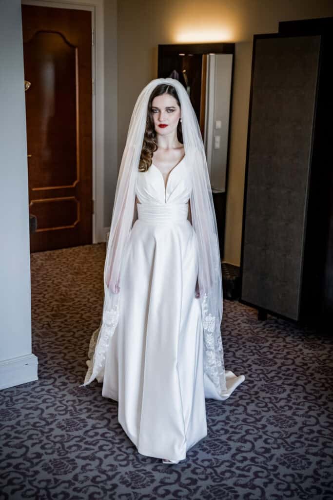Hochzeitsporträt einer Braut in einem eleganten weißen Brautkleid mit Schleier, vor einem modernen Hotelzimmer. Professionelle Hochzeitsfotografie, Fokus auf Stil und Atmosphäre, detaillierte Bildaufnahmen.