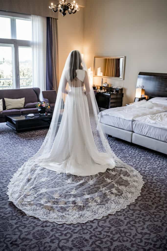 Elegantes Brautkleid mit langem Tüll-Umhang in einem stilvollen Hotelzimmer, bereit für den Hochzeitstag. Hochzeitsfotografie, Innenaufnahme, detailreiche Kleidung, festliche Atmosphäre.