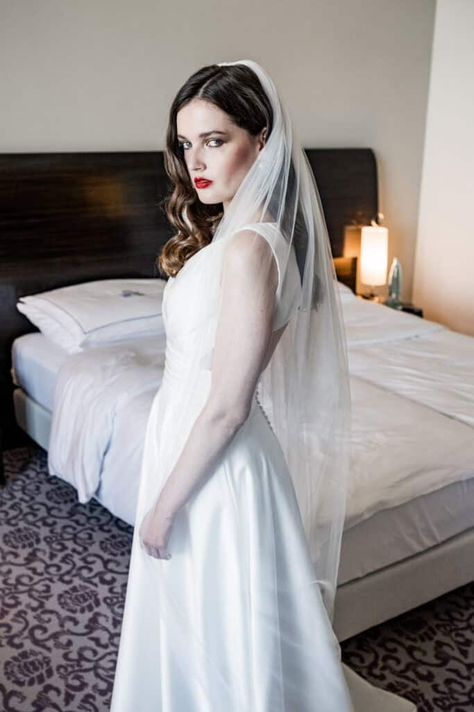 Stilvolle Braut in eleganter Hochzeitskleidung mit Schleier im Hotelzimmer.