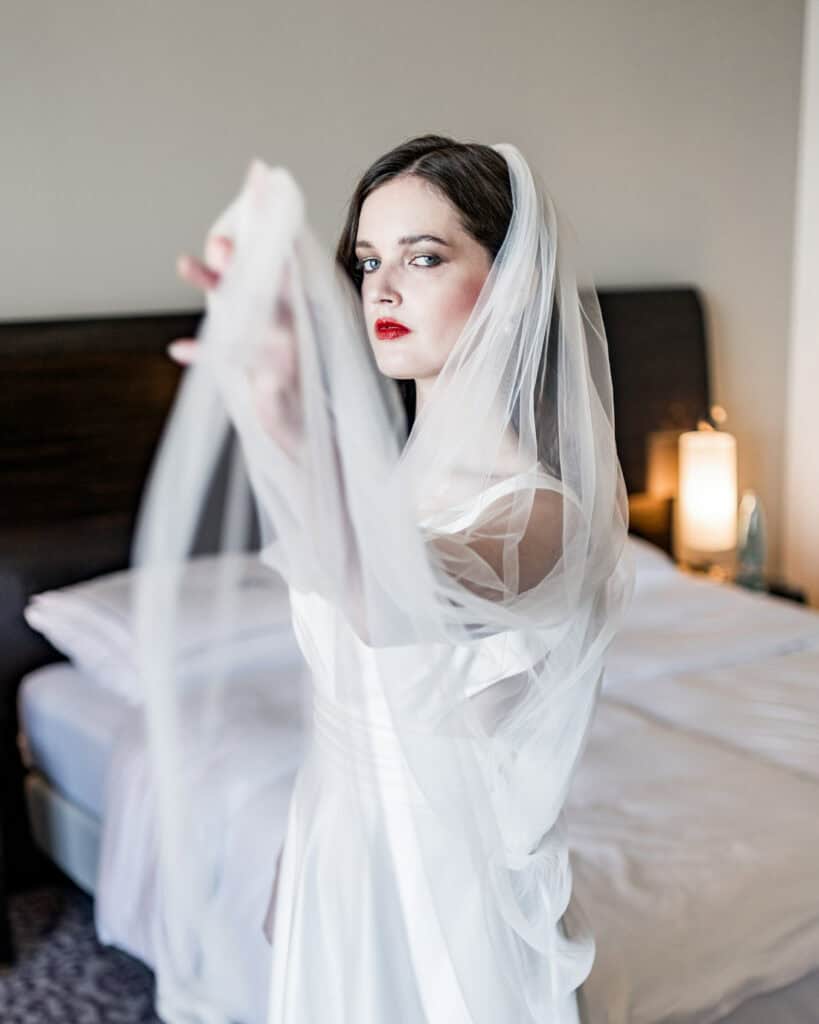 Elegante Braut mit weißem Hochzeitskleid und Schleier im modernen Hotelzimmer, bereit für den besonderen Tag, professionelle Hochzeitsfotografie, emotionale Hochzeitsbilder, Artbild-Fotografie.