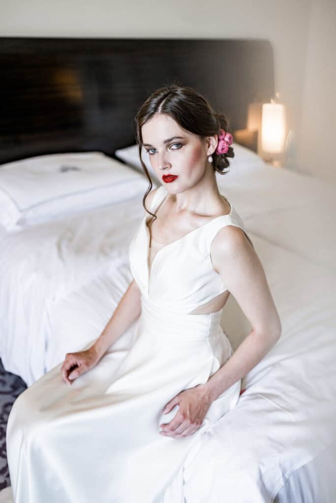 Feine Hochzeitsfotografie einer stilvollen Braut in einem eleganten weißen Kleid, mit rotem Lippenstift, Blumen im Haar und professionellem Make-up, sitzend auf einem Hotelbett in einem modern gestalteten Schlafzimmer.