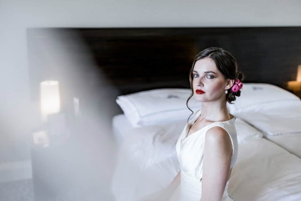 Elegante Braut im weißen Kleid mit roten Lippen und Blumen im Haar vor modernem Schlafzimmer; Hochzeitsfotografie.
