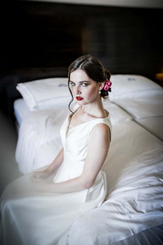 Traumhafte Hochzeitsfotografie einer eleganten Frau in weißem Brautkleid mit rotem Lippenstift und rosa Blumen im Haar, sitzend auf einem Bett in einem stilvollen Schlafzimmer.