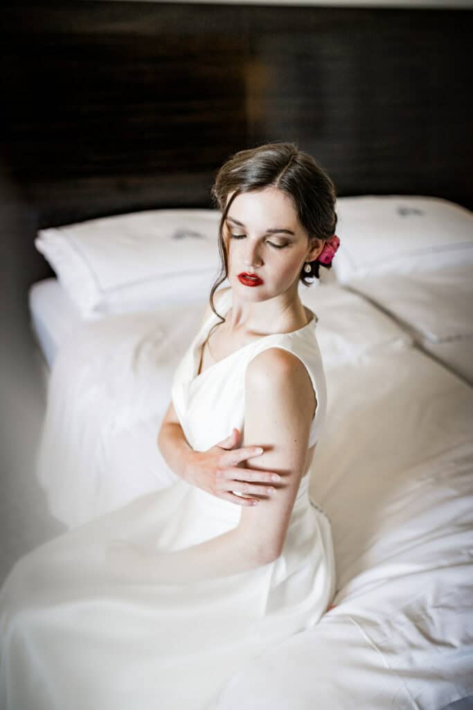 Elegante Frau im weißen Brautkleid sitzt auf einem Bett, romantische Hochzeitsfotografie, emotionale Hochzeitsmotive, professionelle Hochzeitsfotografie, kreative Hochzeitsbilder, Kunstfotografie für besondere Anlässe.