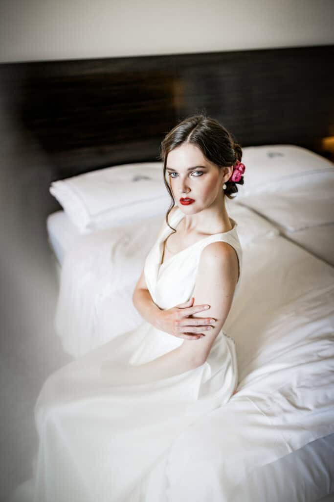 Elegantes Portrait einer jungen Frau in einem weißen Kleid auf Bett, mit rotem Lippenstift und Blumendetail im Haar, bei hoher Qualität, kreative Hochzeits- und Modefotografie.