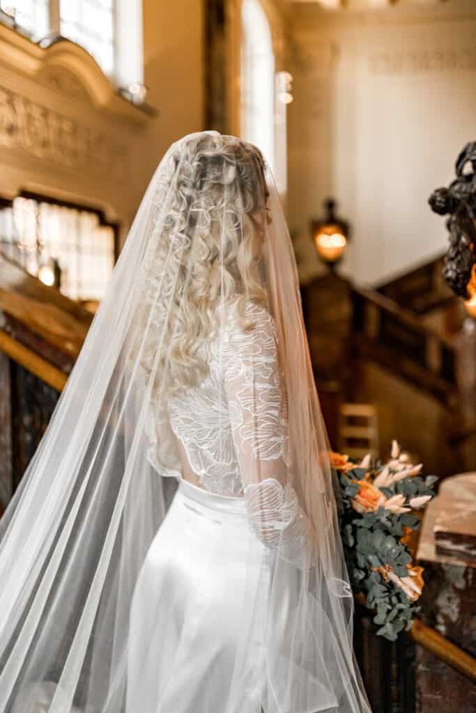 Hochzeitsbraut mit langem Tüllschleier in einem historischen Innenraum, elegante Hochzeitskleidung, romantische Atmosphäre, professionelle Hochzeitsfotografie, kunstvolle Porträtaufnahme.